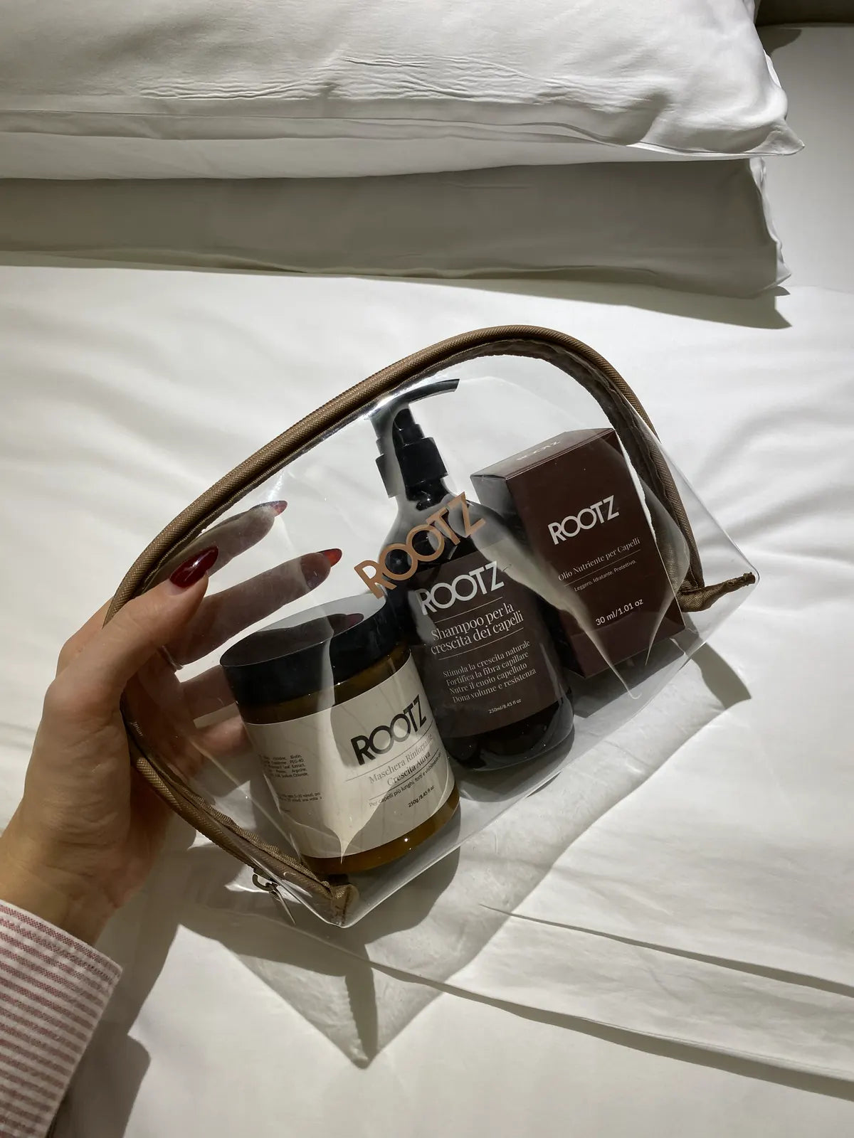Una mano levanta el neceser transparente con el champú, la mascarilla y el aceite dentro, sobre una cama deshecha
