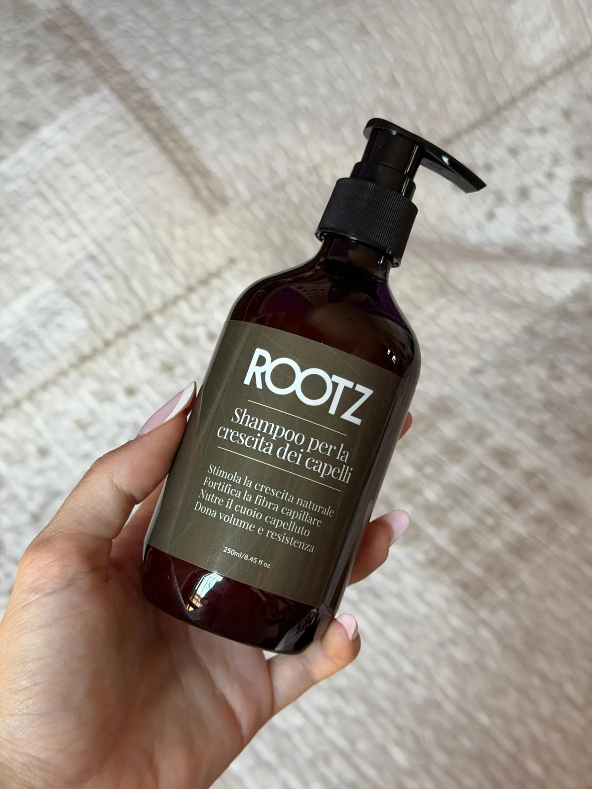 Mano che regge il flacone da 250 ml dello shampoo ROOTZ sopra un tappeto chiaro