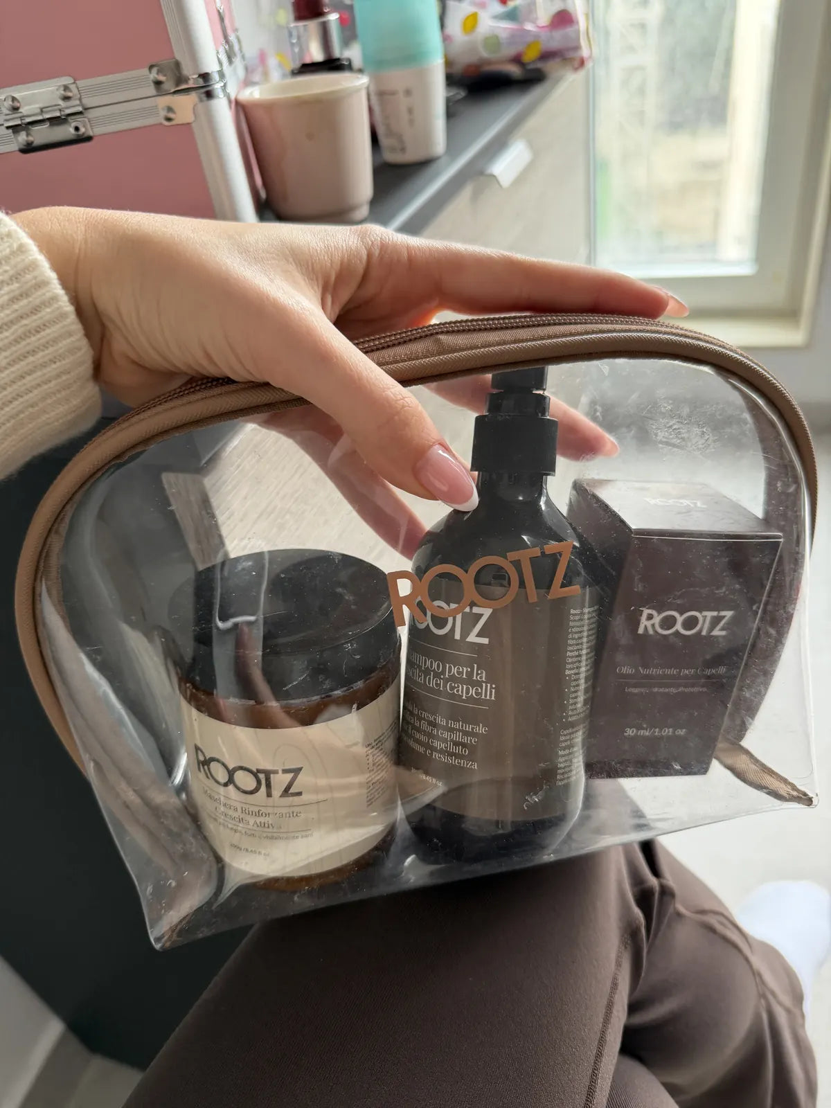 El neceser transparente con los productos ROOTZ apoyado en el regazo, en el baño